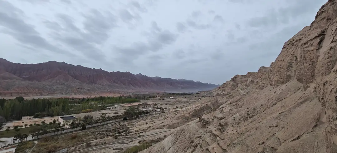 Looking past the cliffs of the Kizil Caves to green groves and distant rust-red ridges