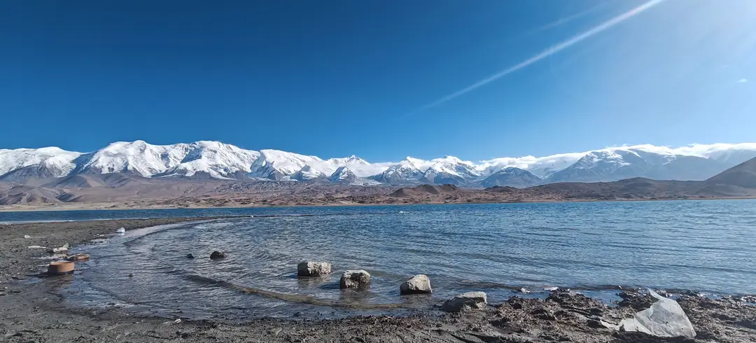 Ripples wash around shoreline rocks on Karakul Lake, with snow-covered peaks beyond